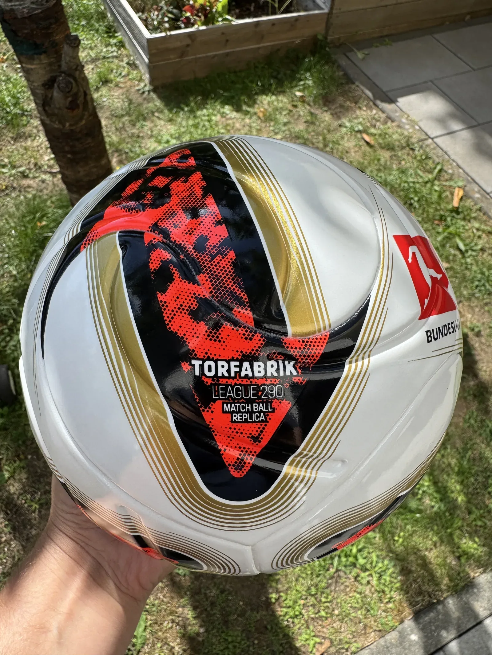 Weiß-schwarz-rot-goldener adidas-Fußball wird im Garten in einer Hand gehalten. Lesbar sind TORFABRIK, LEAGUE 290 und MATCH BALL REPLICA.