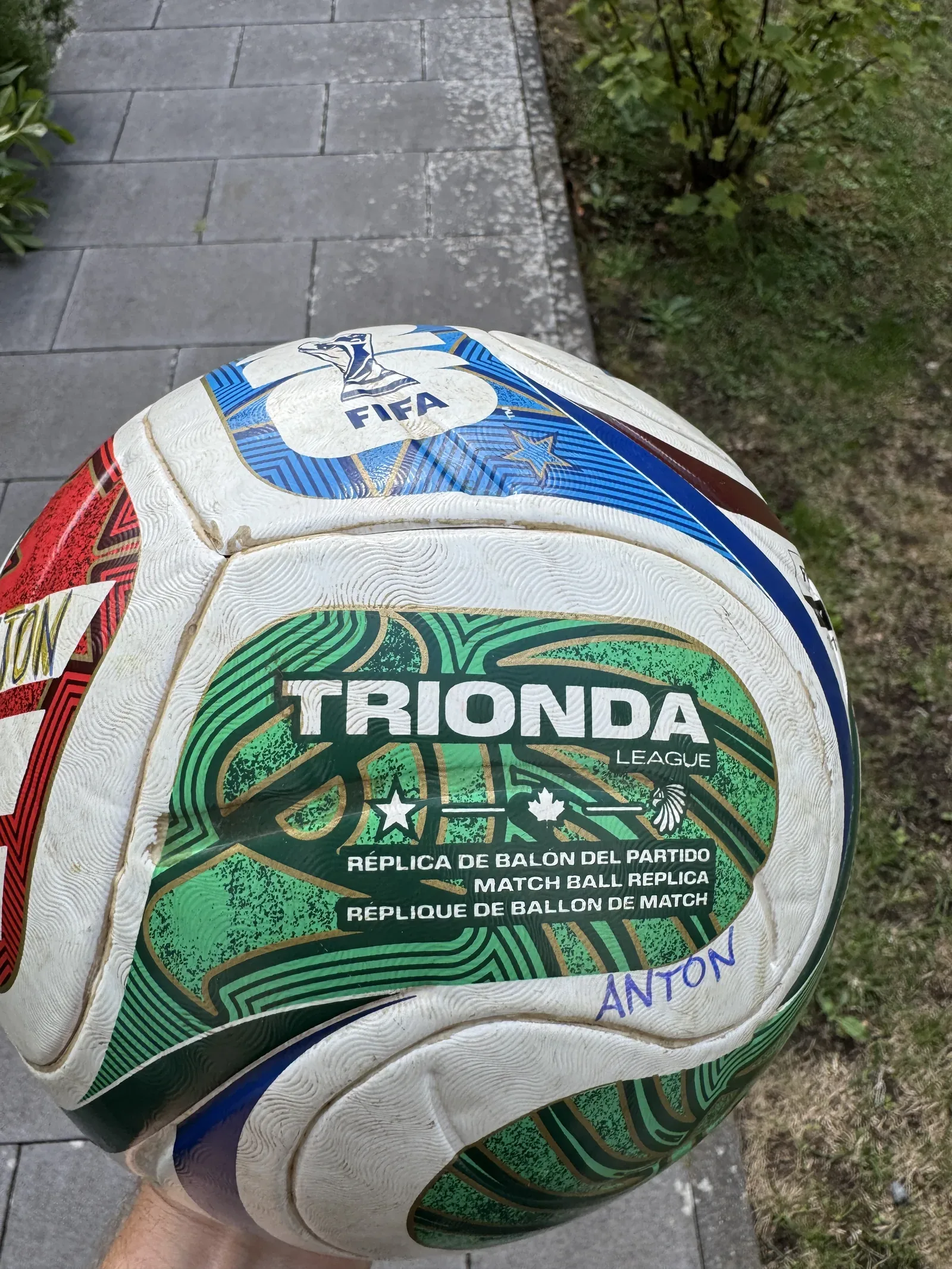 Weißer Fußball mit grünen, roten und blauen Bahnen in einer Hand, mit dem Aufdruck Trionda League und Match Ball Replica