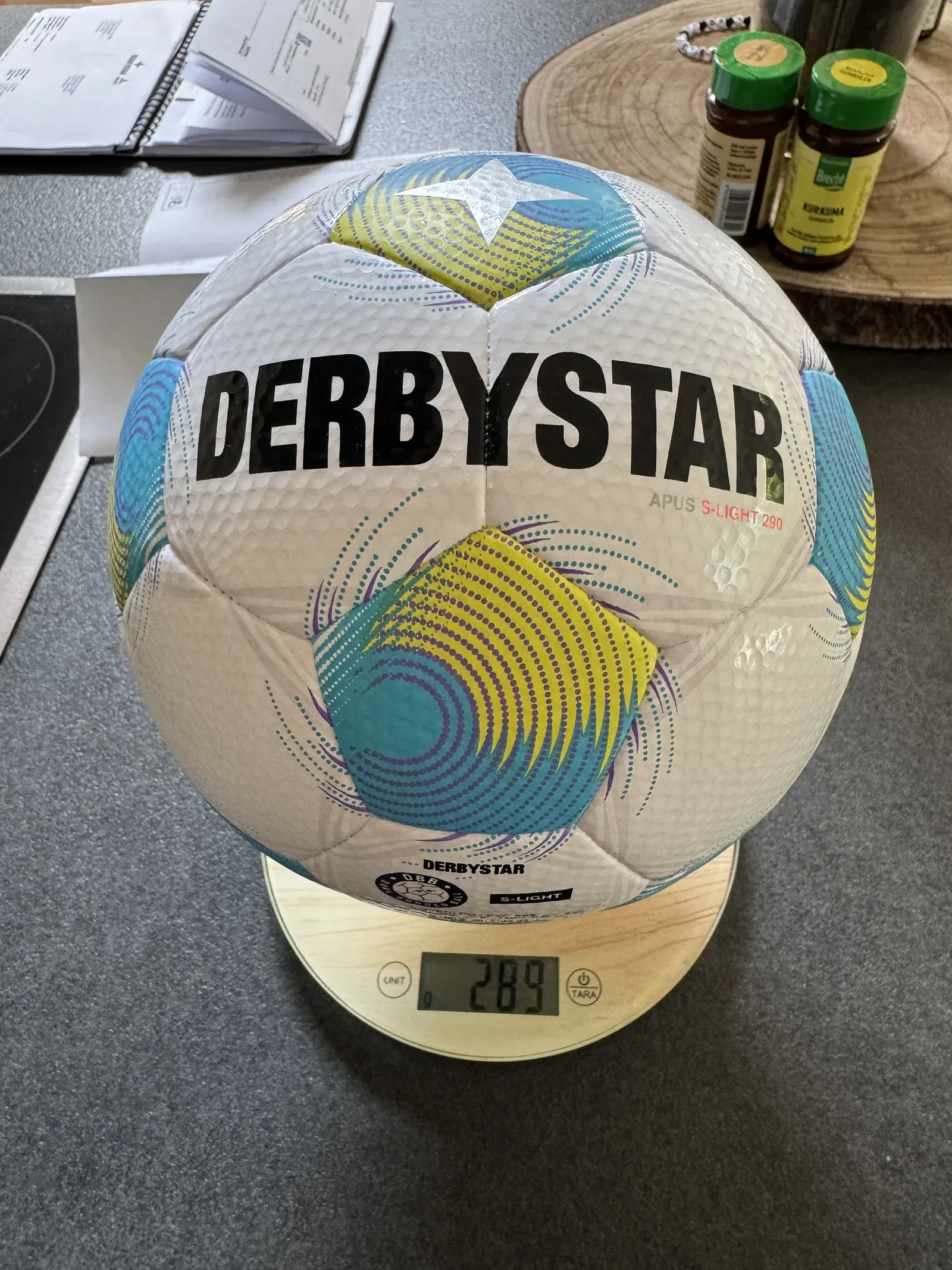 Weißer Fußball mit türkis-gelb-violettem Muster auf einer Küchenwaage, die 289 anzeigt. Auf dem Ball lesbar: DERBYSTAR, APUS S-LIGHT 290.