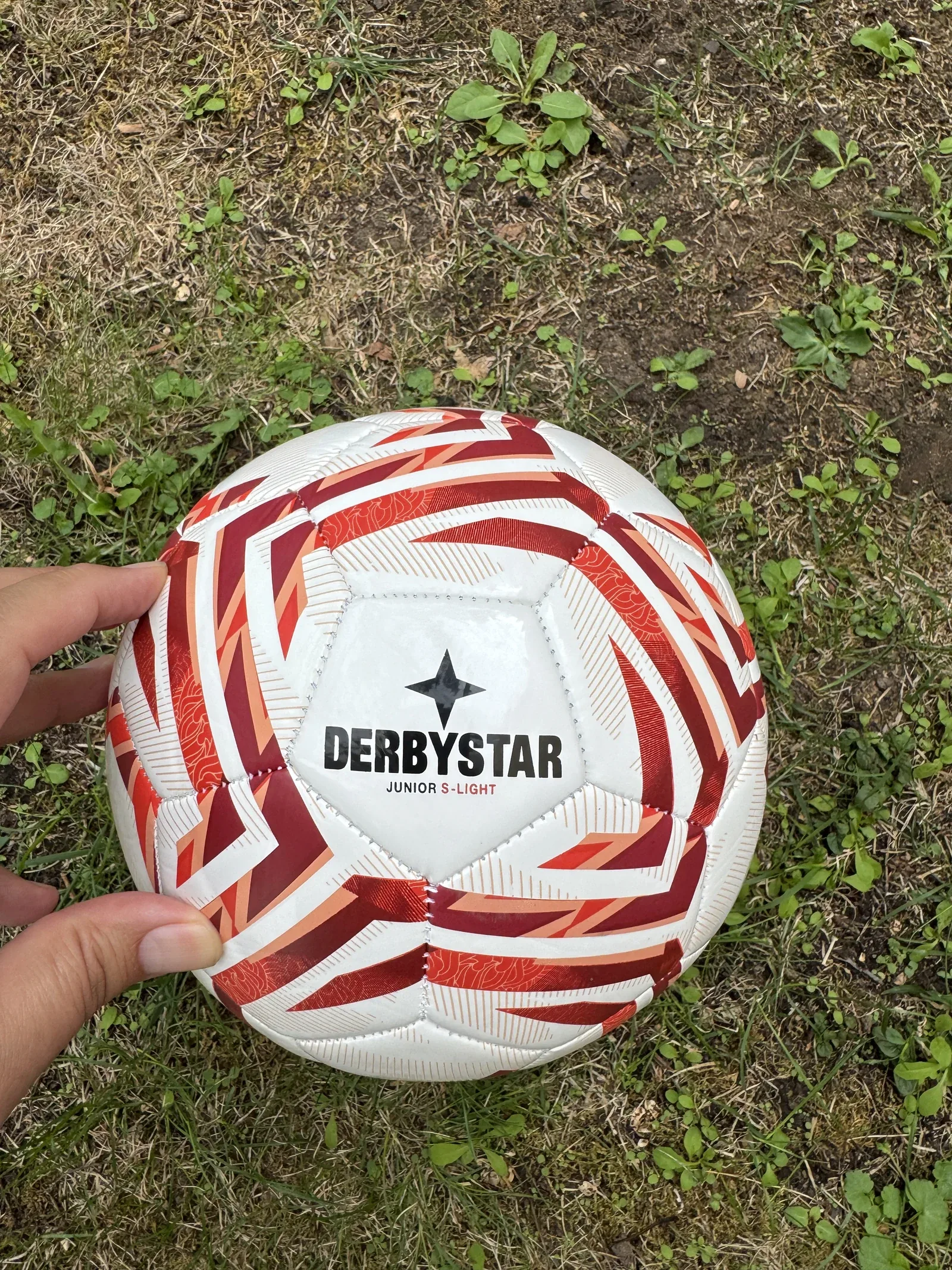 Rot-weißer Fußball im Gras, von einer Hand am linken Bildrand gehalten. Auf dem oberen Feld der vierzackige Derbystar-Stern, darunter DERBYSTAR und klein JUNIOR S-LIGHT