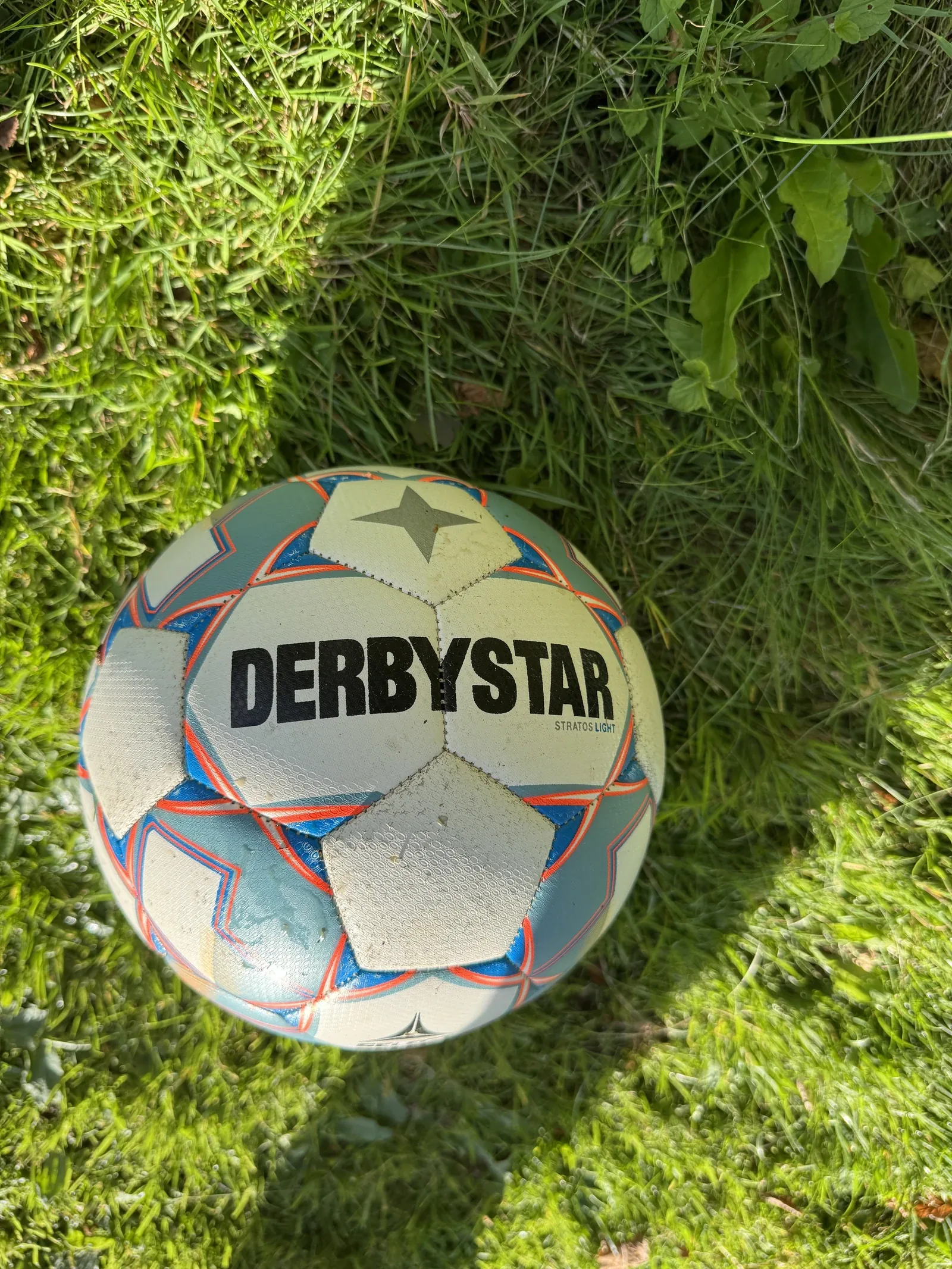 Silber-blau-oranger Fußball im hohen Gras. Auf der hellen Fläche steht DERBYSTAR und daneben klein Stratos Light, darüber ein Stern.
