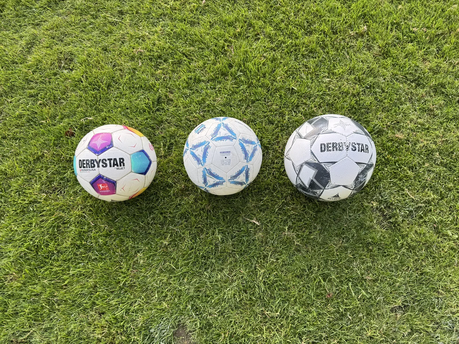 Drei Fußbälle nebeneinander im Gras, von links nach rechts größer werdend: ein bunter Derbystar Bundesliga Brillant Replica, ein weiß-blauer Sport-Thieme Core Kids mit dem Aufdruck SIZE 4 und ein abgespielter schwarz-weißer Derbystar
